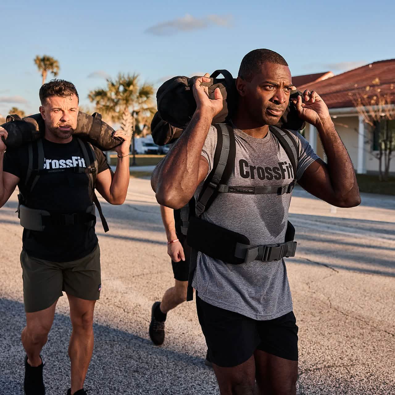 Two men undertake an outdoor CrossFit workout, carrying substantial weights on their shoulders. They sport GORUCK's Padded Hip Belts as they train on a paved surface, surrounded by palm trees and a building looming in the background.