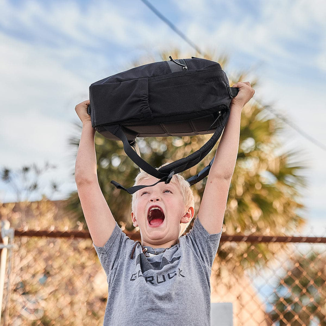 A child joyfully lifts a durable GORUCK KR1 book bag overhead outdoors, with trees and a fence in the background.