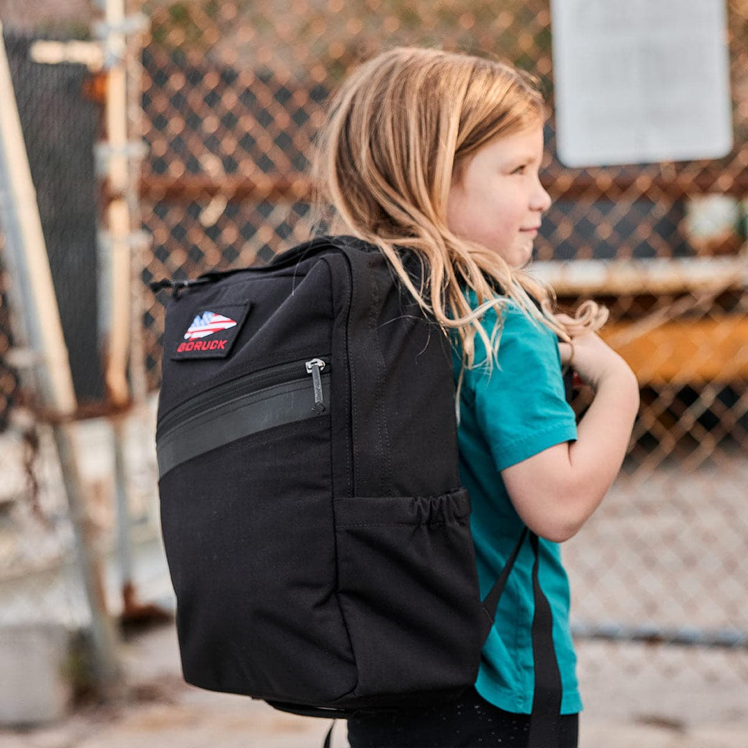 A child with long hair wearing a teal shirt carries a GORUCK KR1 book bag near a chain-link fence.