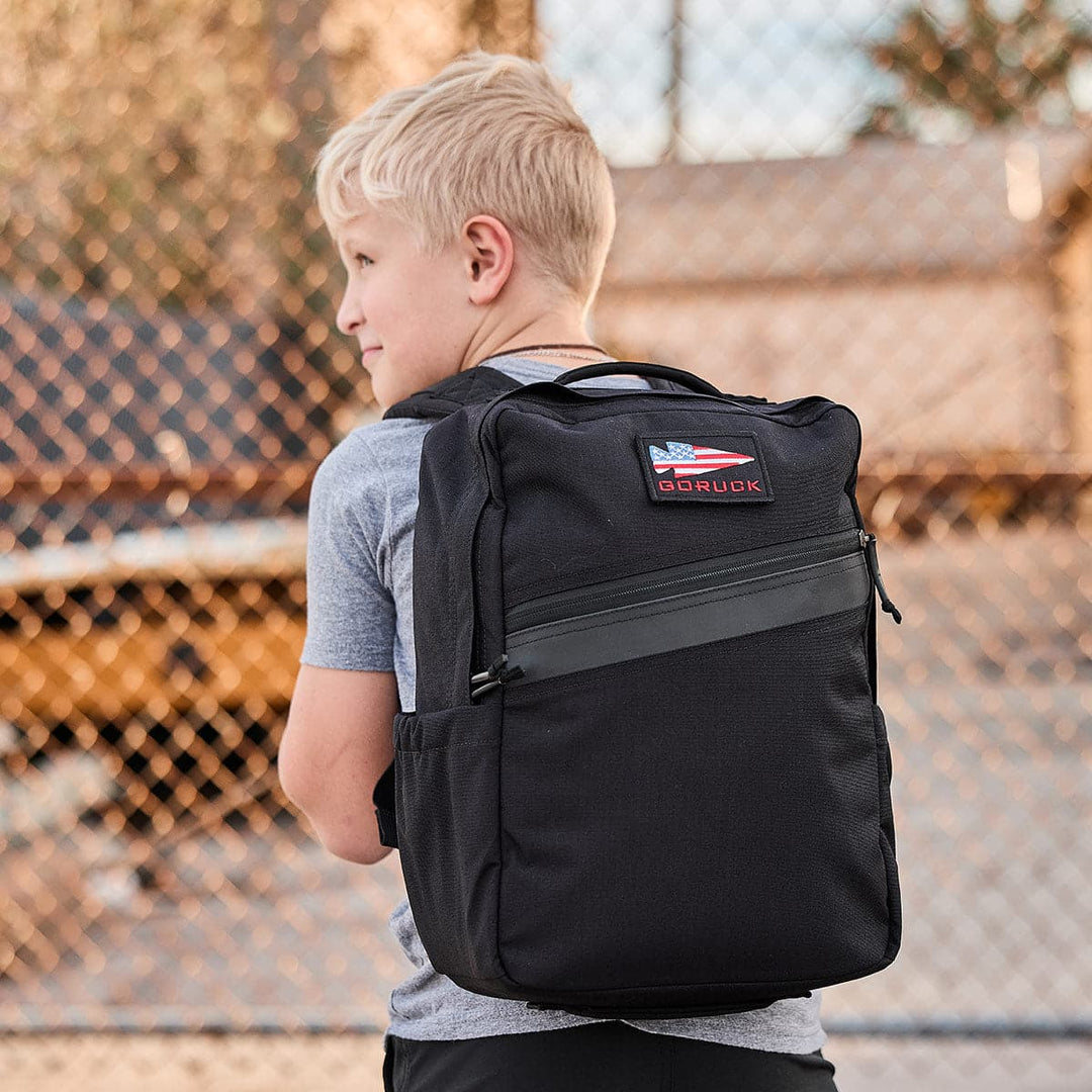 A blonde boy wearing a gray shirt holds a GORUCK KR1 black durable book bag near a chain-link fence.