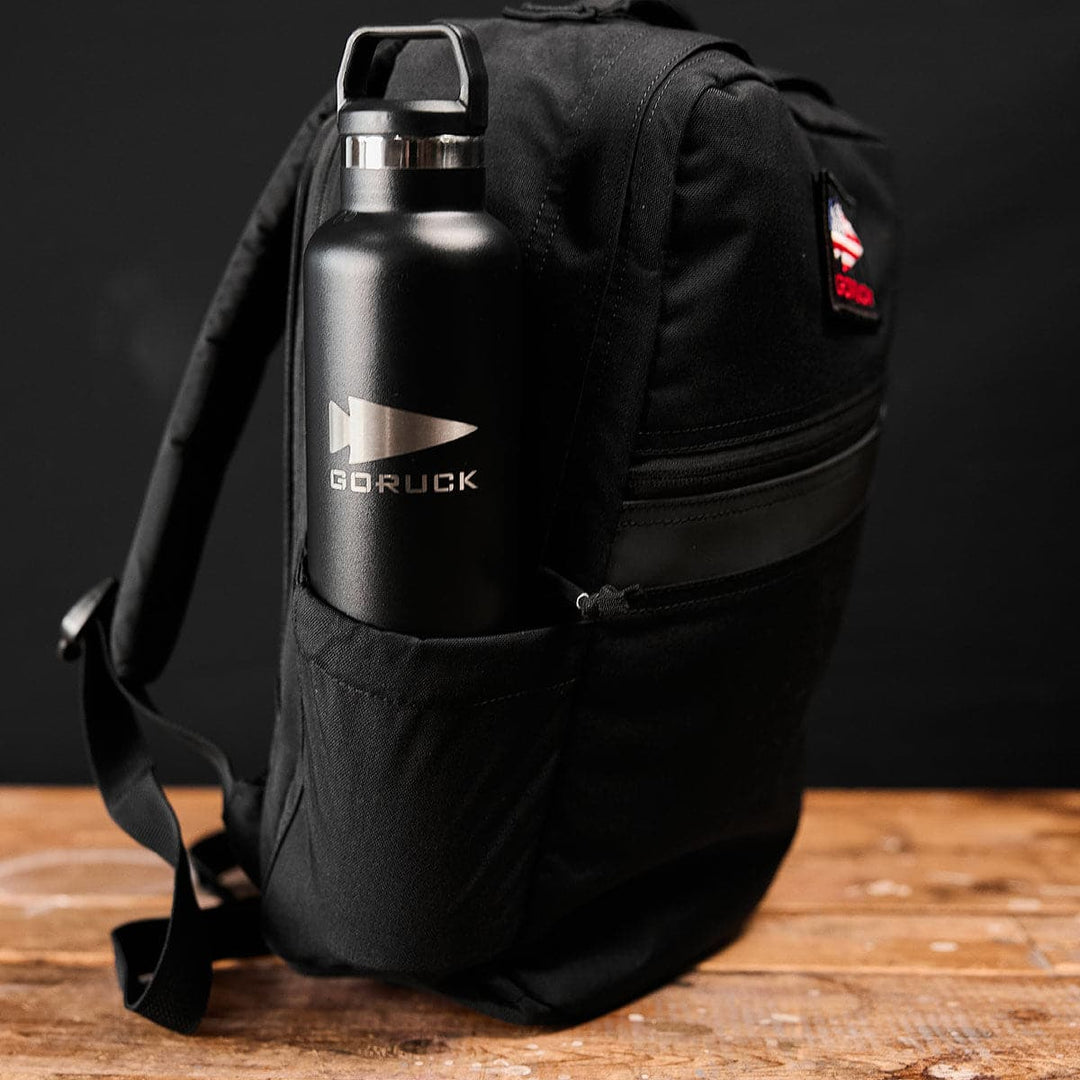 A durable KR1 book bag rests on a wooden surface, highlighting a GORUCK bottle securely nestled in the side pocket.