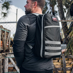 Man in a gray long-sleeve shirt wearing a black backpack stands outdoors near a chain-link fence and palm trees.