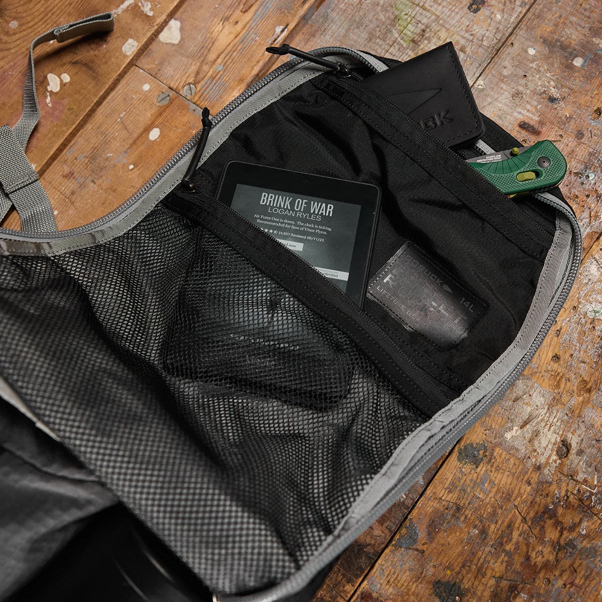 An open backpack on a wooden floor, with an e-reader, wallet, knife, and card in its inner pockets.