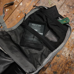 An open backpack on a wooden floor, with an e-reader, wallet, knife, and card in its inner pockets.