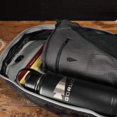 A backpack contains a GORUCK water bottle, a book, and a gray pouch on a wooden surface.