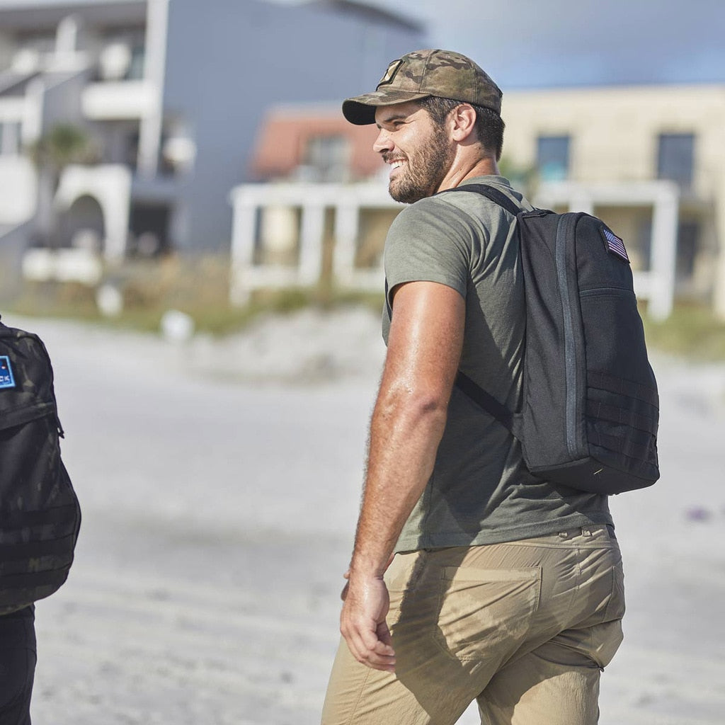 Man in a t-shirt and cap walking outside, wearing a backpack, with buildings in the background.