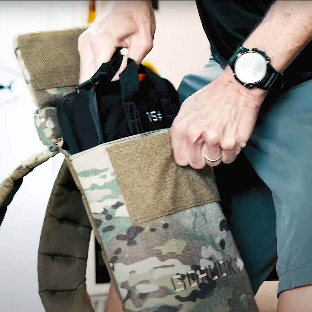 A person is carefully placing a black bag into a camouflage-patterned backpack, equipped with the convenient GORUCK Sand Ruck Plates. They're wearing a stylish watch and ring, ready for some serious rucking.