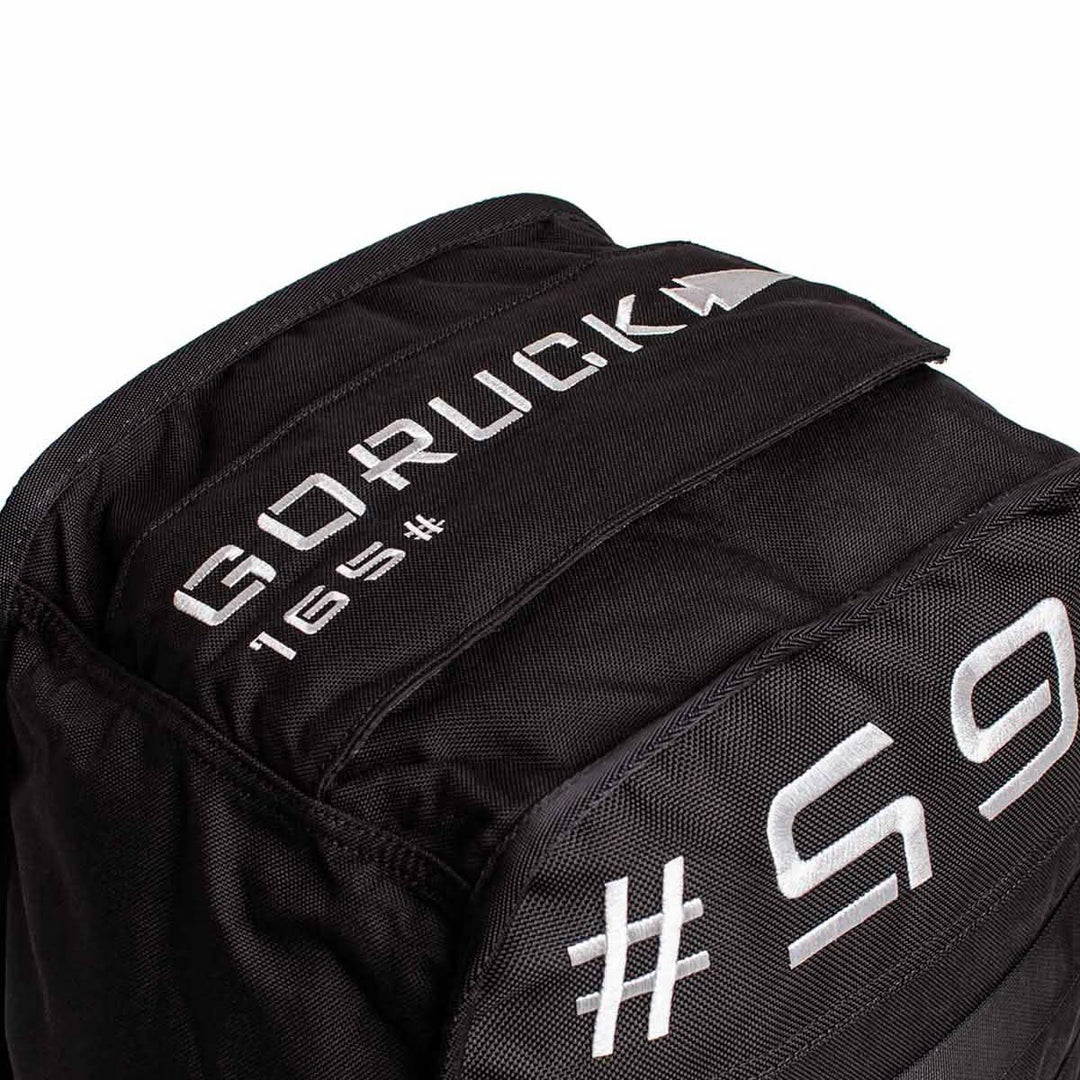 A close-up of a black Sand Tombstones by GORUCK showcases white embroidered text, including GORUCK, 165, and partial text 165 on the fabric. Its rugged design features a multilayer closure system, making it perfect for loaded carries and inspired by the durability of the Husafell Sandbag.