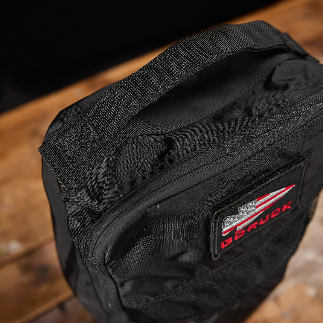 Close-up of a black **Shoe Bag - Ripstop ROBIC** by **GORUCK** on a wooden surface, featuring a logo with red and white elements. The bag is made from water-resistant material, making it perfect for any weather.