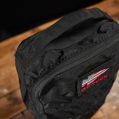 Close-up of a black **Shoe Bag - Ripstop ROBIC** by **GORUCK** on a wooden surface, featuring a logo with red and white elements. The bag is made from water-resistant material, making it perfect for any weather.