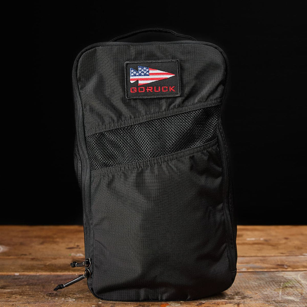 A durable black GORUCK shoe bag made from Ripstop ROBIC with a USA flag patch sits on a wooden surface, making it ideal for any journey.