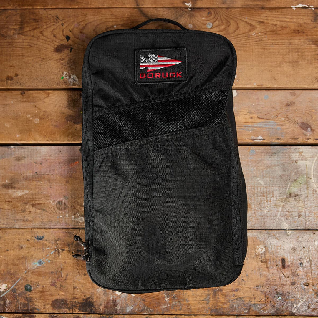 The Shoe Bag - Ripstop ROBIC from GORUCK, showcasing a sleek design, sits on a wooden floor. This durable and water-resistant accessory combines practicality with style, making it ideal for any adventure.