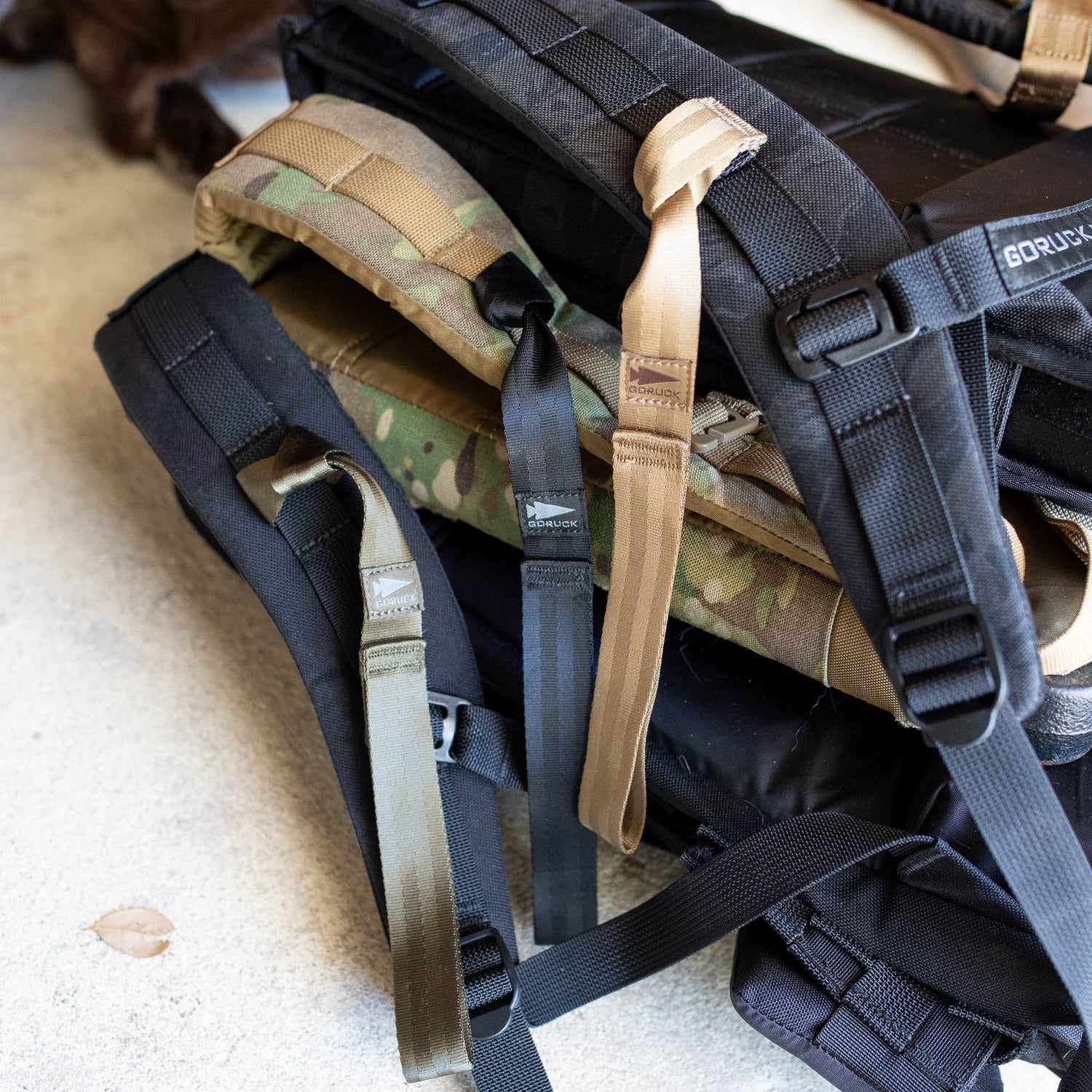 Three backpacks with black, green, and tan straps stacked on concrete, each equipped with T-REX Hand Straps for comfort and hand swelling relief during long rucks.