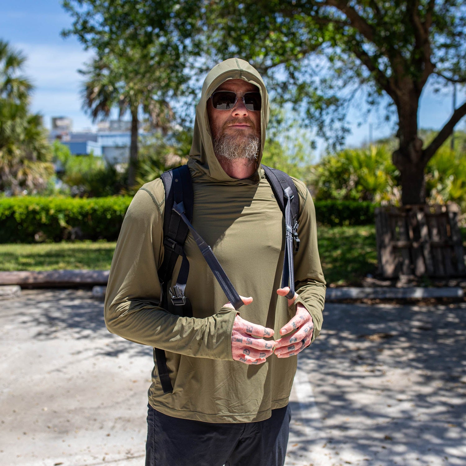 A man in sunglasses and a hooded shirt stands outdoors with a backpack, using T-REX Hand Straps—a must-have rucking accessory—amid trees and sunlight.