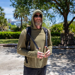 A man in sunglasses and a hooded shirt stands outdoors with a backpack, using T-REX Hand Straps—a must-have rucking accessory—amid trees and sunlight.