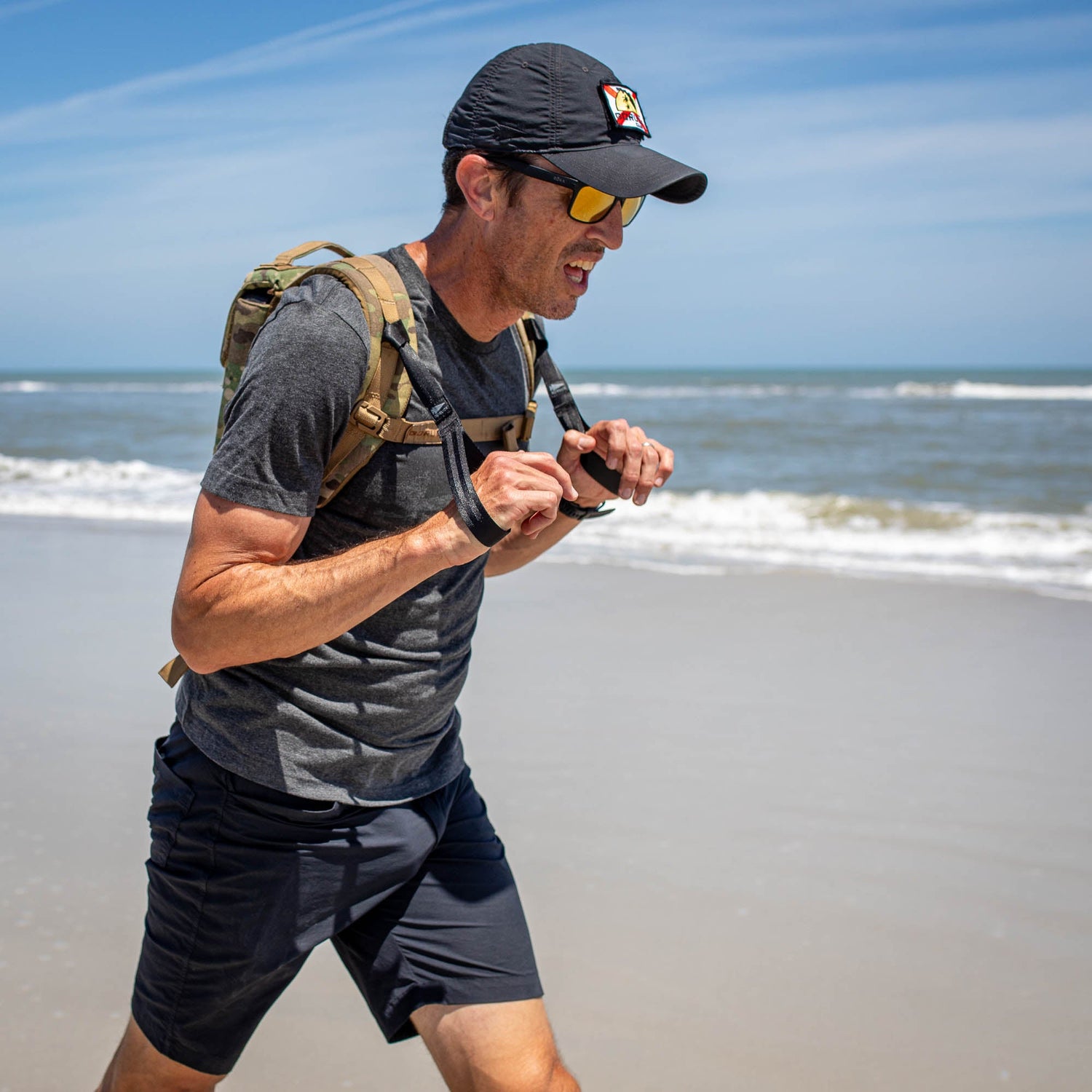 A man with a cap and backpack, wearing T-REX Hand Straps for hand swelling relief, walks briskly on a sunny beach by the ocean.