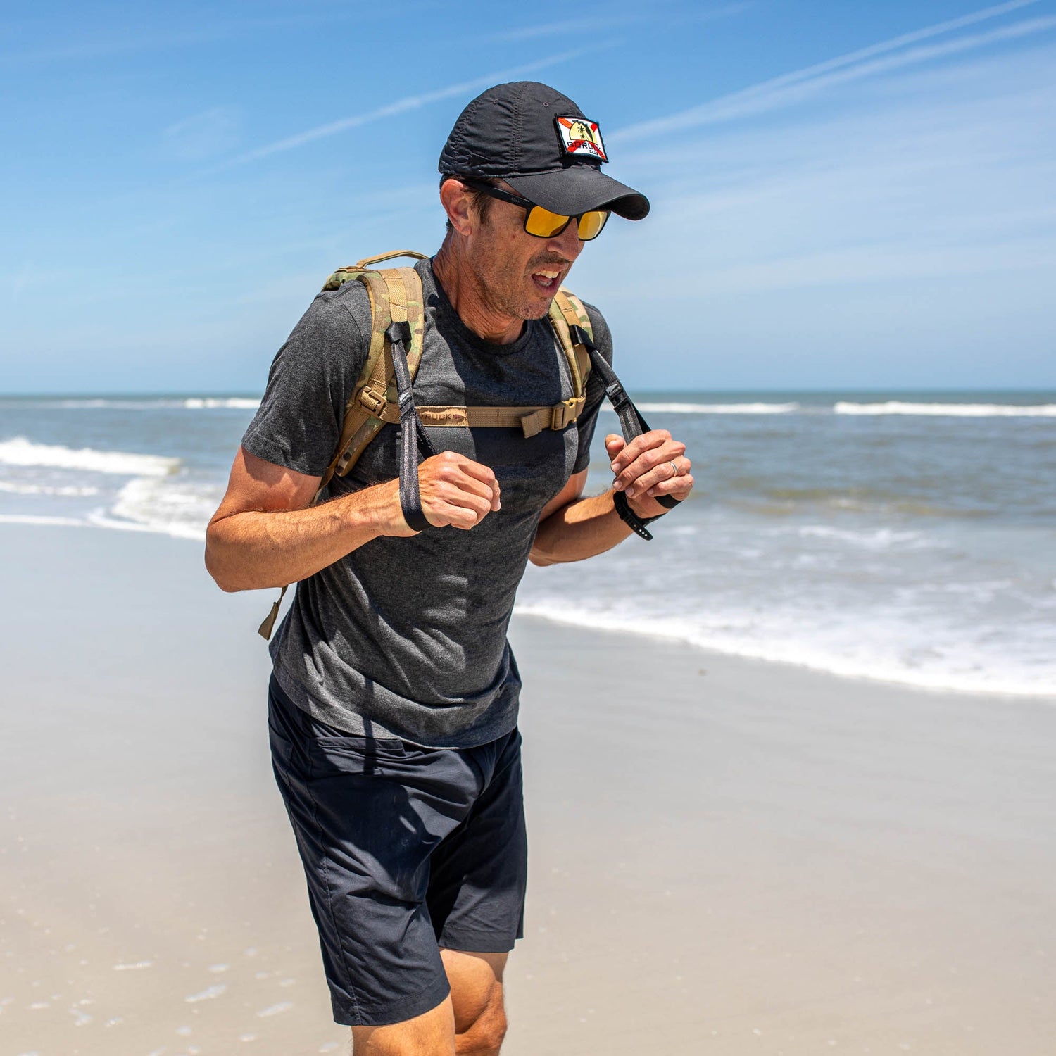 A man jogs on a sandy beach near the ocean under a clear blue sky, wearing sunglasses and a backpack with T-REX Hand Straps—his preferred rucking accessory.
