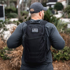 A man with the Bullet Ruck - Classic -15L backpack, dressed in dark clothing, walks outdoors surrounded by greenery and palm trees.