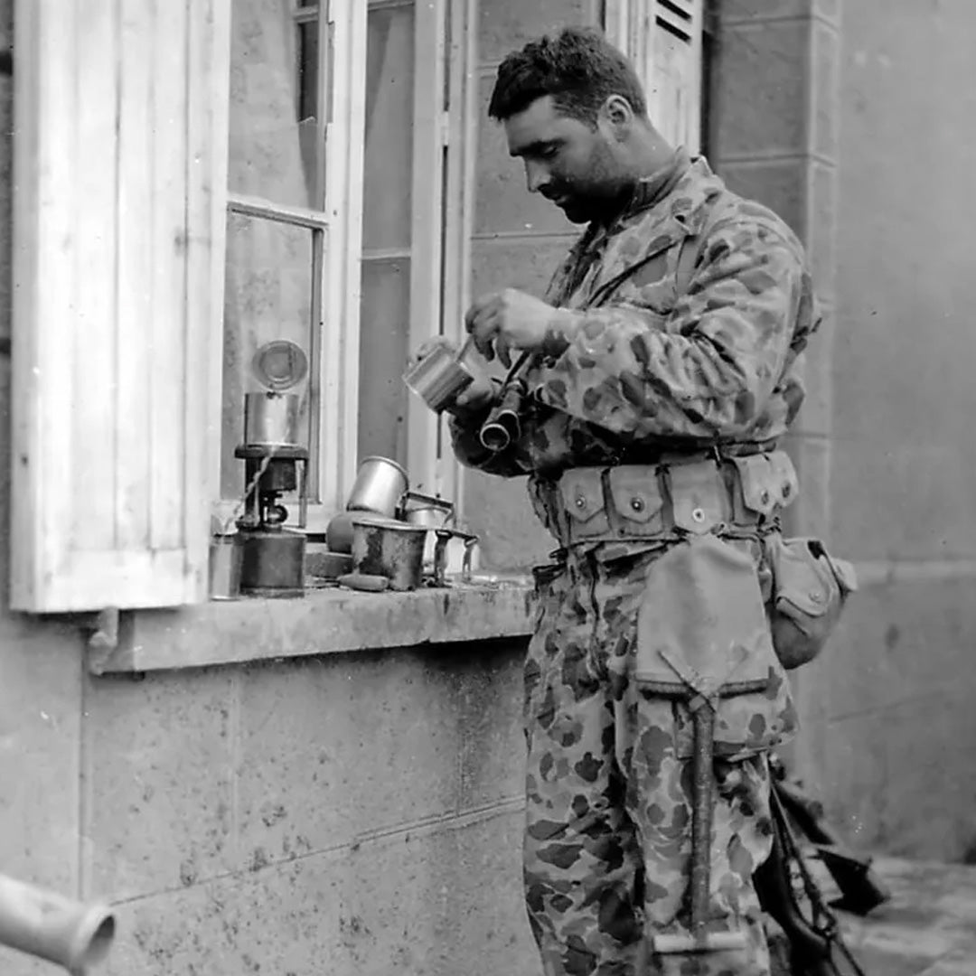 A soldier in camouflage uniform makes coffee on a windowsill using camping equipment and mugs.