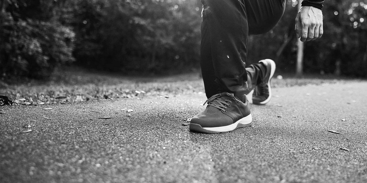 A person in athletic shoes kneels on an outdoor path, surrounded by trees in the background.