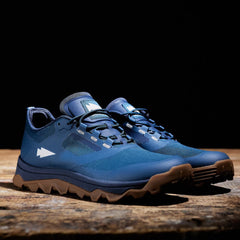 The Mackall trail running shoes by GORUCK in Legion Blue + Gum rest on a wooden surface against a dark background, their robust brown soles revealing the all-terrain challenges they are designed to overcome.