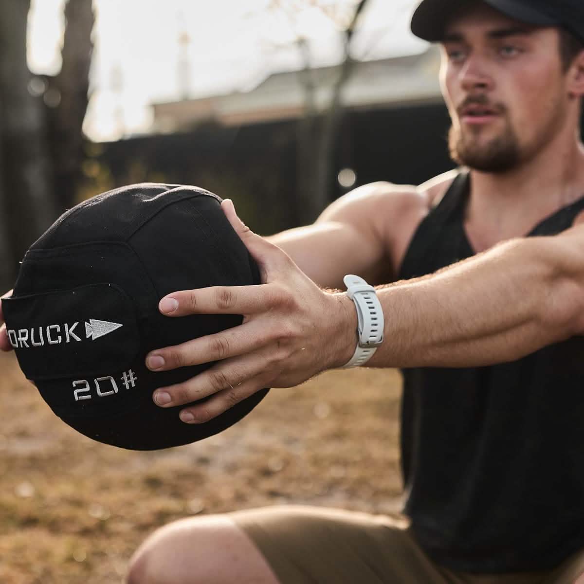 A man is exercising outdoors in a park, holding a 20-pound GORUCK Sand Medicine Ball to enhance core stability.