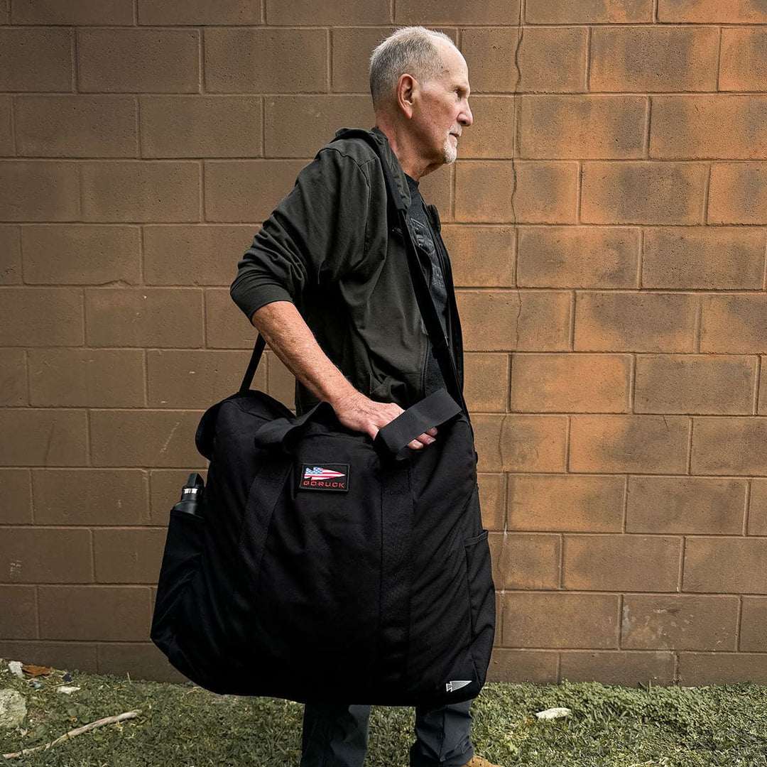 A man stands against a brick wall, carrying the Kit Bag - 84L by GORUCK over his shoulder.