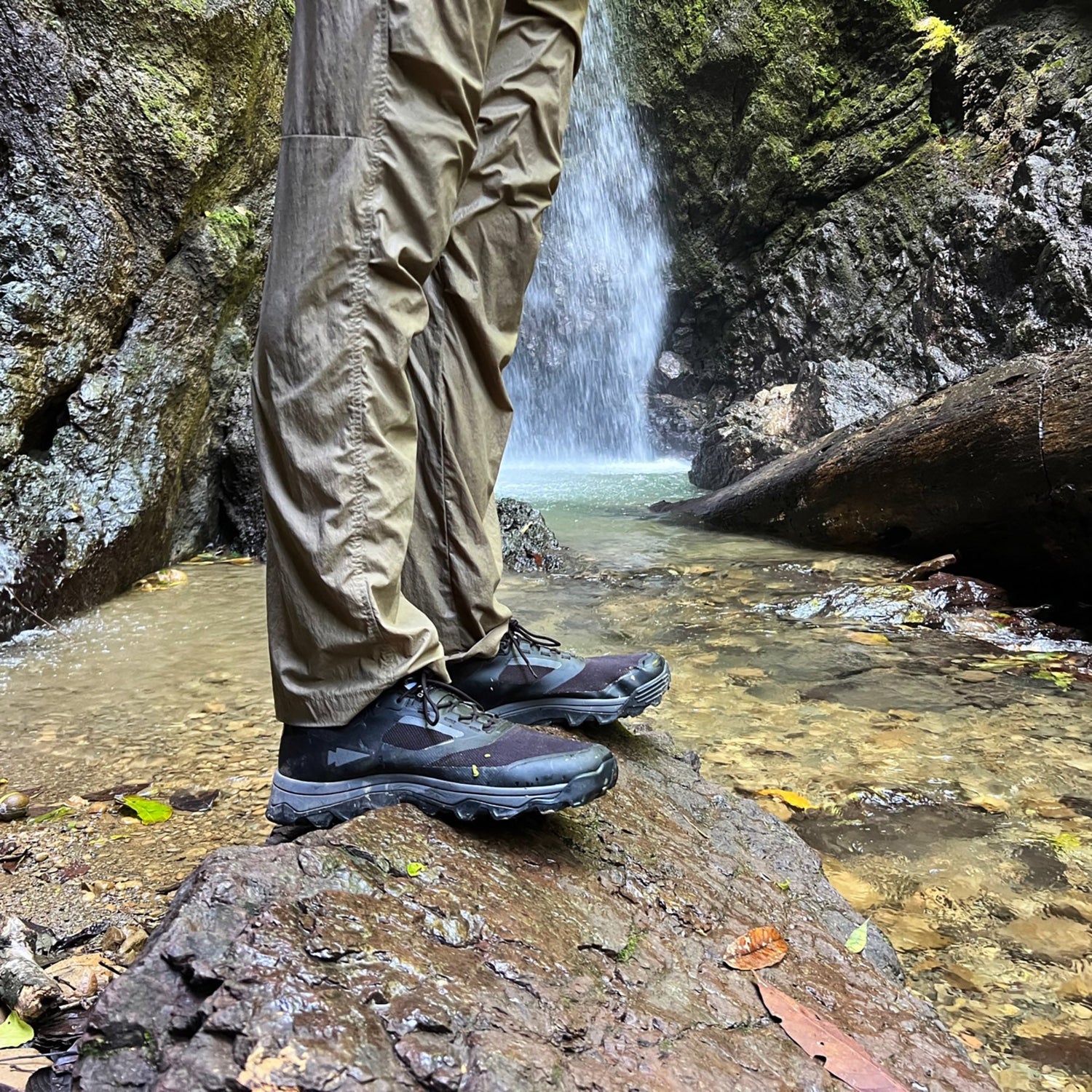 Person wearing hiking boots stands on a rock by a stream and waterfall in a forested area.