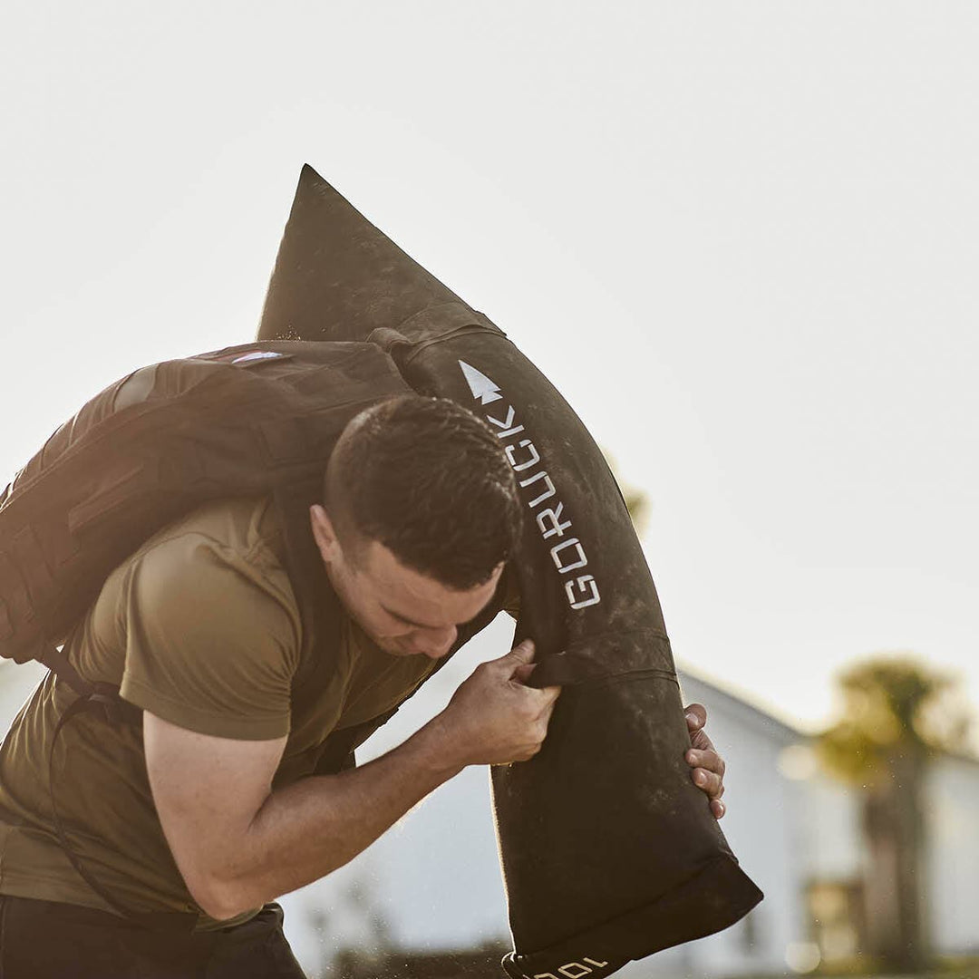 A man in a green t-shirt is training outdoors with the GORUCK Simple Training Sandbags, demonstrating the durability and versatility of his gear, akin to a home gym setup.