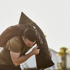 A man in a green t-shirt is training outdoors with the GORUCK Simple Training Sandbags, demonstrating the durability and versatility of his gear, akin to a home gym setup.