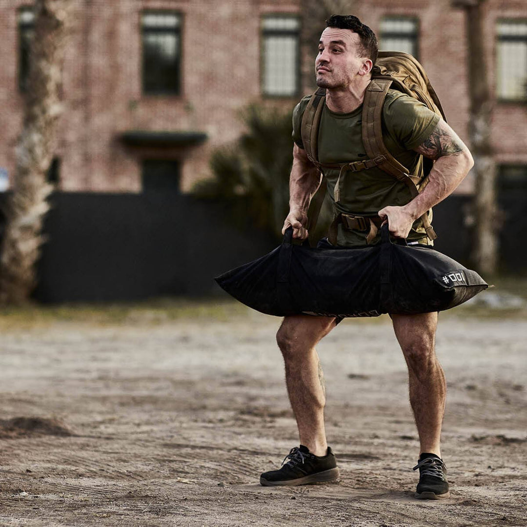 A man with a GORUCK backpack and athletic clothing stands confidently in front of a brick building, embodying his dedication to fitness, while carrying a large bag filled with Simple Training Sandbags.