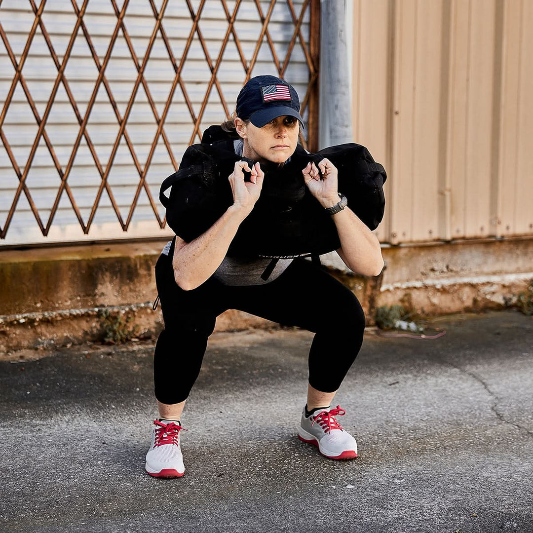Someone is squatting outdoors with a weighted bag on their shoulders, wearing athletic gear and a cap, demonstrating the art of rucking with the Ruck Clubs Starter Kit - Light by GORUCK.