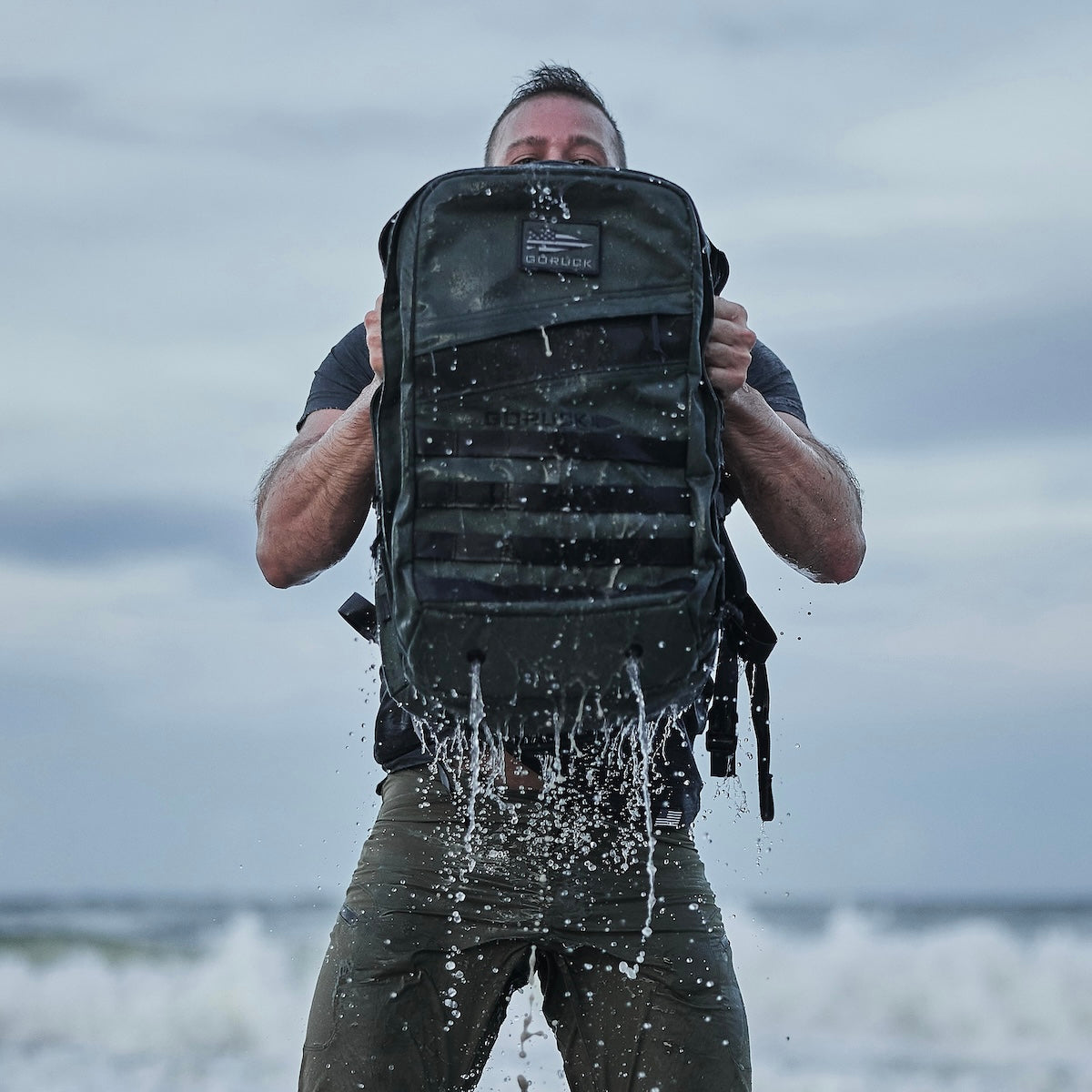 Man holding a wet backpack in front of him as water pours out, standing outdoors with cloudy sky behind.
