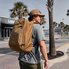 Man wearing a tan GORUCK backpack outdoors, palm trees and shops in background