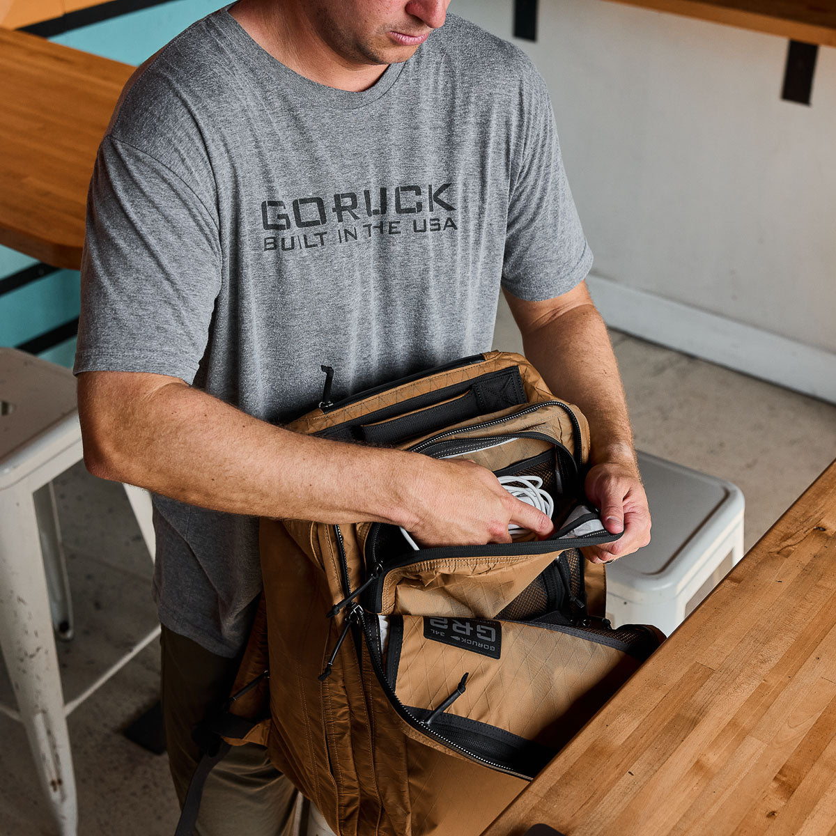 Man wearing GORUCK t-shirt organizes gear in tan GORUCK GR2 backpack indoors
