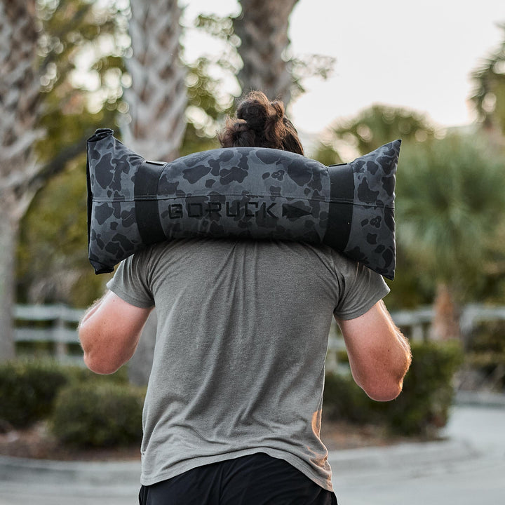 A person carries a black camo GORUCK Simple Training Sandbag outdoors near palm trees, highlighting a versatile workout tool for any home gym, backed by the Scars Lifetime Guarantee.