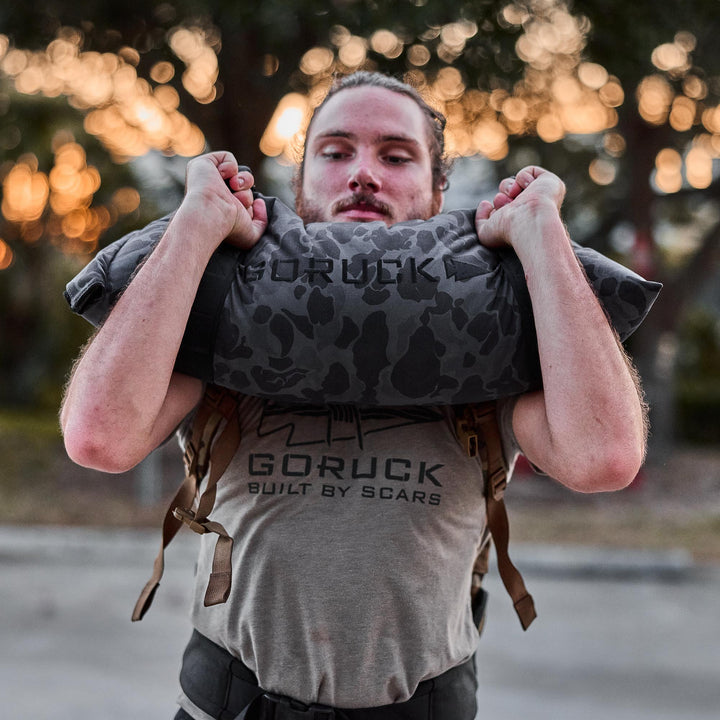A person lifts a GORUCK Simple Training Sandbag outdoors at sunset, wearing a "GORUCK Built by Scars" t-shirt—demonstrating grit and the Scars Lifetime Guarantee.