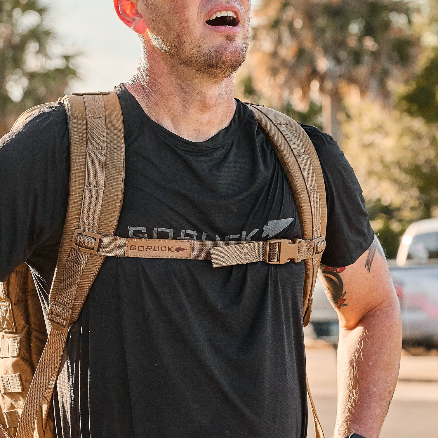 A person wearing a black GORUCK shirt and backpack equipped with the GORUCK Training Sternum Strap, standing outdoors with trees and cars in the background.
