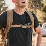 A person wearing a black GORUCK shirt and backpack equipped with the GORUCK Training Sternum Strap, standing outdoors with trees and cars in the background.