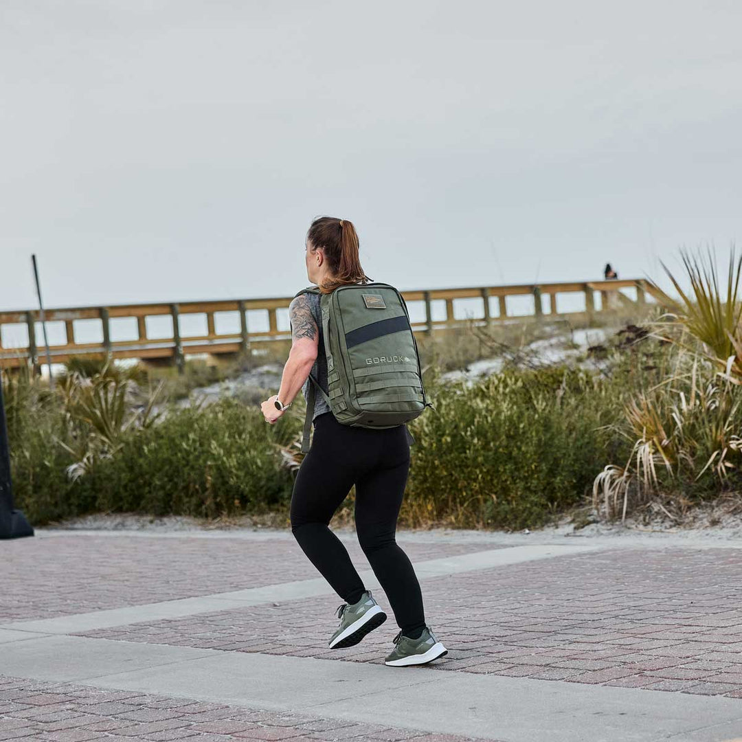 A person is rucking along a paved path near a boardwalk, wearing black clothes and carrying a green GORUCK Rucker 4.0 backpack loaded with Ruck Plates.