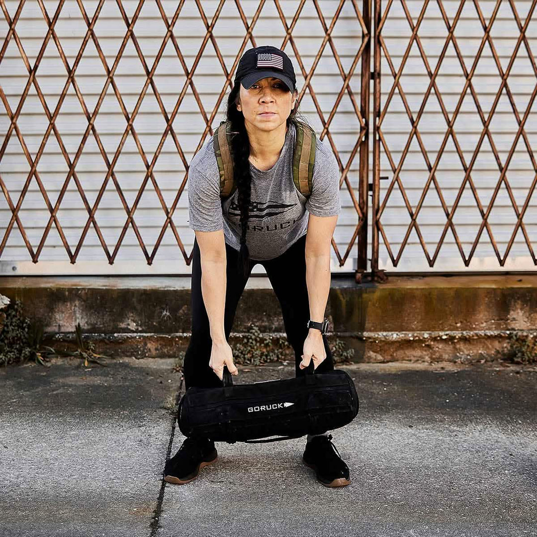 A woman in athletic wear lifts a GORUCK Sandbag Kit (w/ Filler Bag), made from durable 1000D CORDURA, outdoors in front of a metal gate.