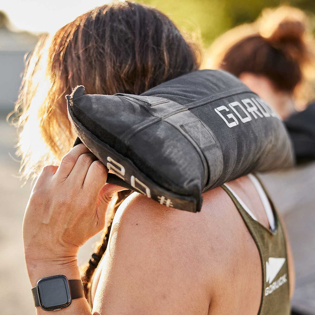 A person is training with a GORUCK Simple Training Sandbag slung over their shoulder in a sunny outdoor setting, utilizing the heavy-duty handles for better grip and control.