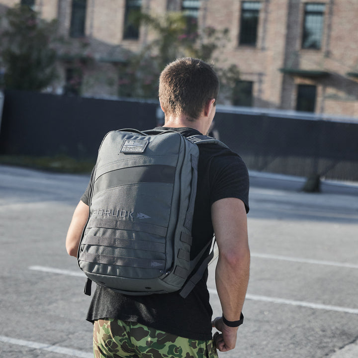 A man wearing camouflage pants and carrying the GORUCK Rucker 4.0 backpack walks outdoors near a brick building.