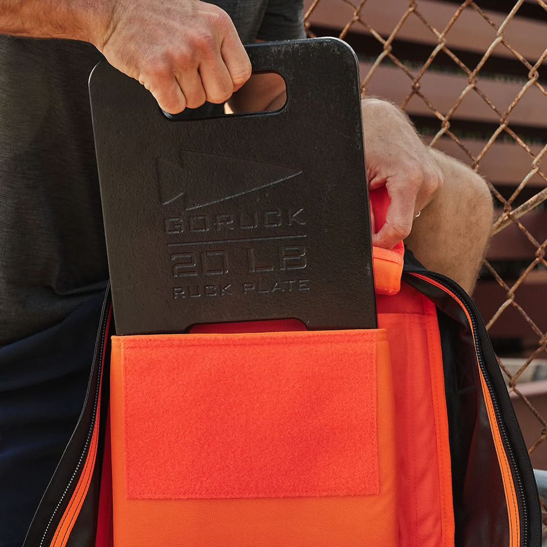 An individual carefully inserts the Ruck Clubs Starter Kit - Light from GORUCK, weighing 20 lbs, into an orange backpack, gearing up for their rucking adventure.