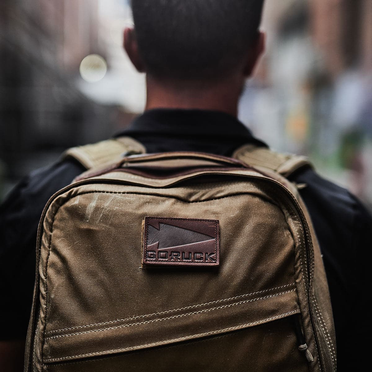 A person wearing a GORUCK GR1 USA - Heritage backpack adorned with patches strolls through a blurred urban setting, showcasing its rugged charm.