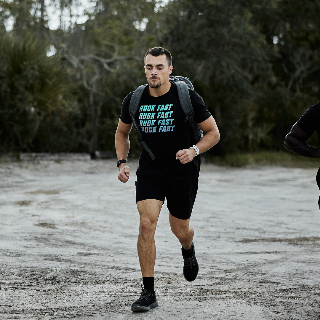 A man runs outside on a dirt path wearing a black t-shirt, shorts, and carrying the GORUCK Rucker 4.0—ideal for rucking with GORUCK Ruck Plates.