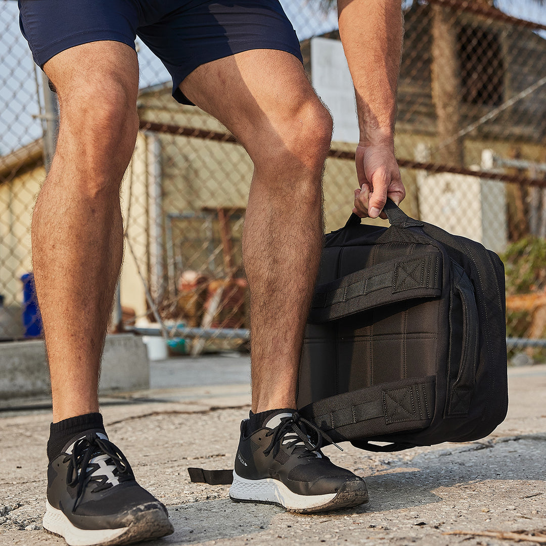 Wearing athletic shoes and shorts, a person lifts the GORUCK Rucker 4.0 black rucksack near a chain-link fence, ready for some serious rucking outdoors.