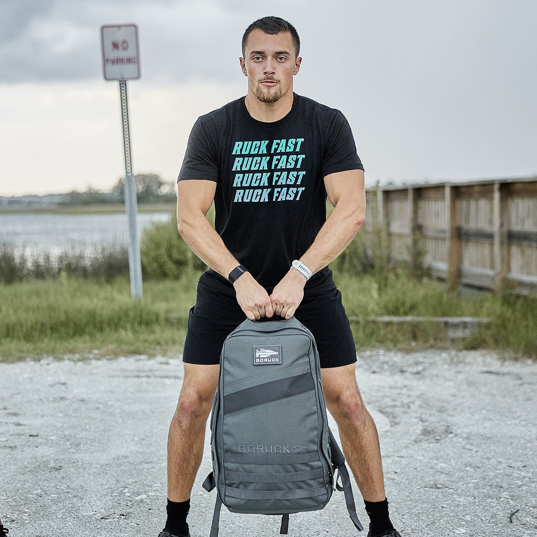 A man in a "Ruck Fast" shirt stands outdoors holding the GORUCK Rucker 4.0 gray rucksack by its top handle, ready to start rucking.