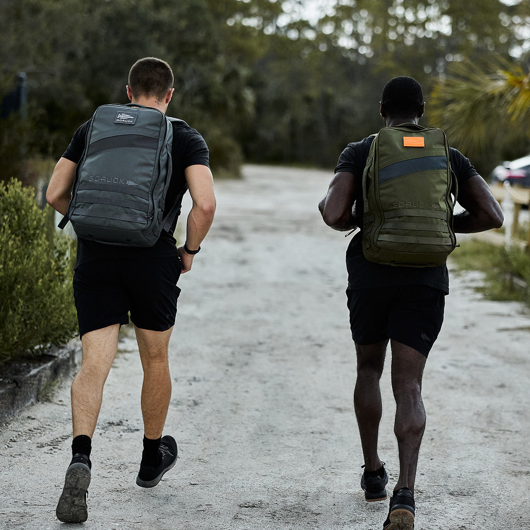 Two men with rucksacks walk or jog down a gravel path surrounded by greenery, enjoying a morning of rucking with the GORUCK Rucker 4.0.