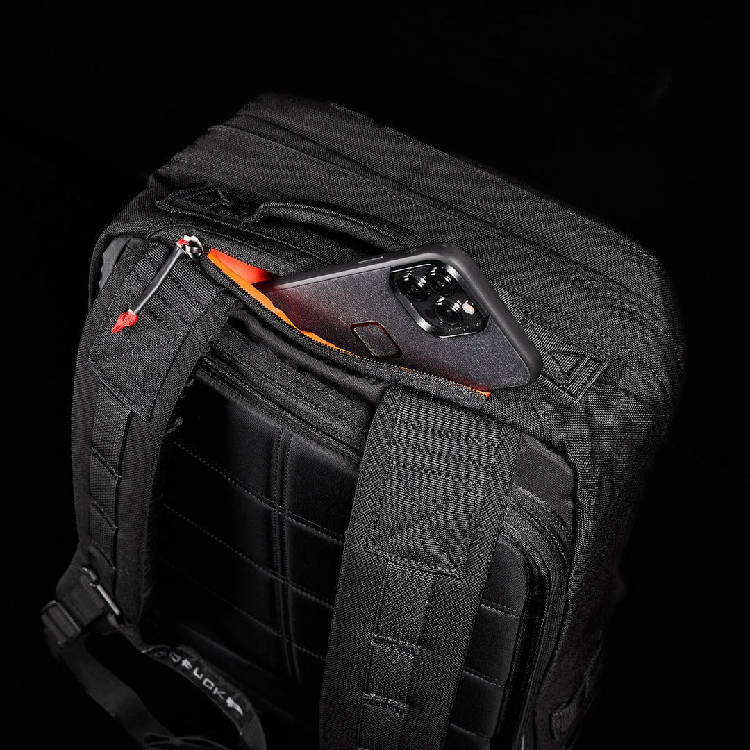 A smartphone partially peeks from the top pocket of a black GORUCK GR2 backpack against a stark black background, showcasing its durable design inspired by Special Forces medical packs.